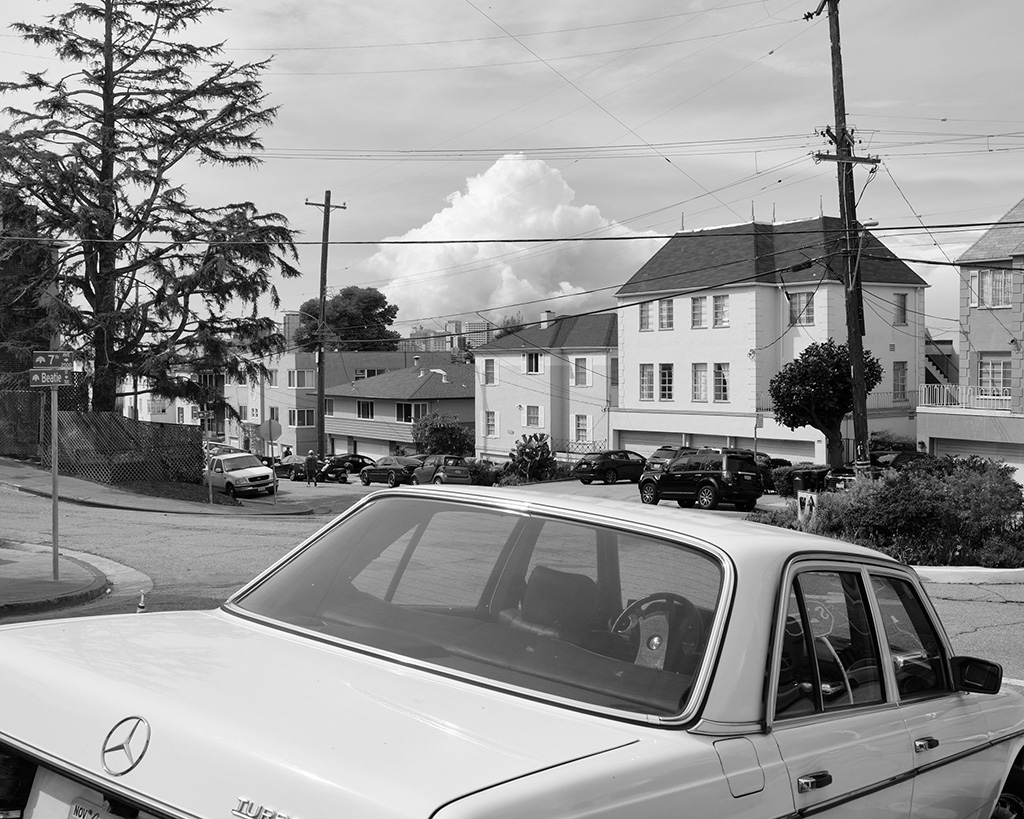 A black and white photograph of a roundabout in Oakland, California, with a car in the foreground and a cloud that looks like a mountain in the distance