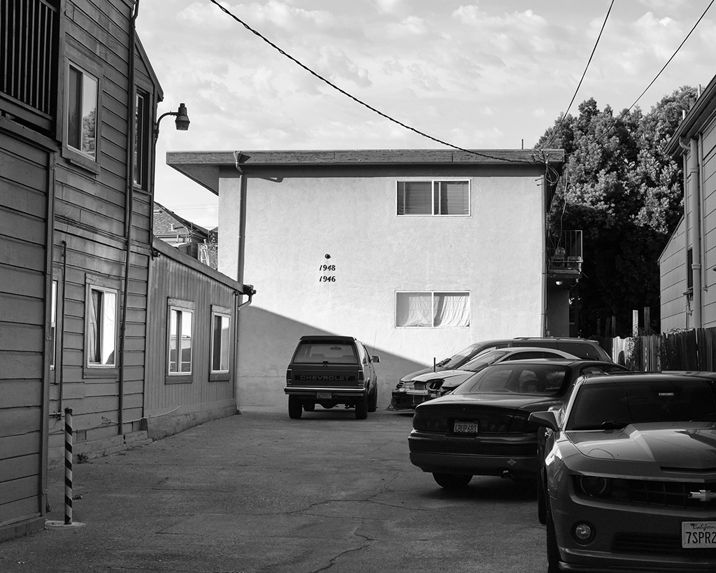 A black and white photograph of a parking area between apartment buildings in Oakland, California