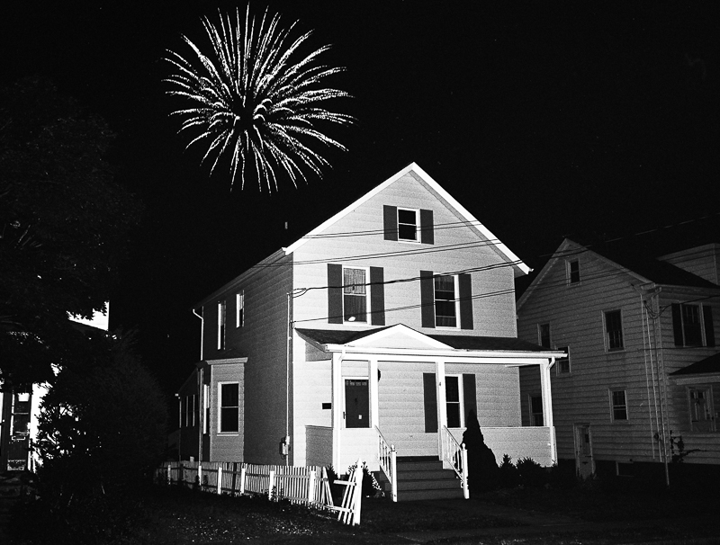 A black and white photograph of a suburban home, at night, with a firework exploding behind it